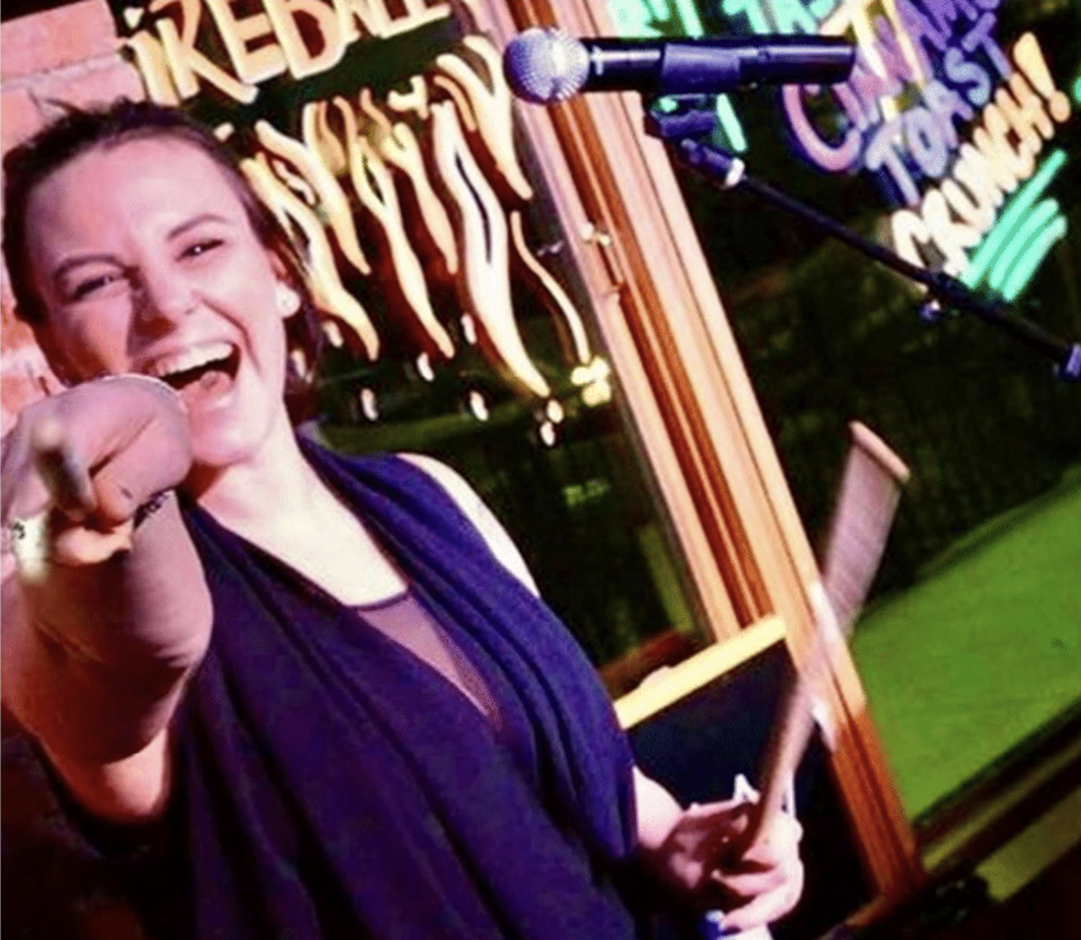 A woman laughing and pointing at the camera while holding a stick, with neon signs in the background.