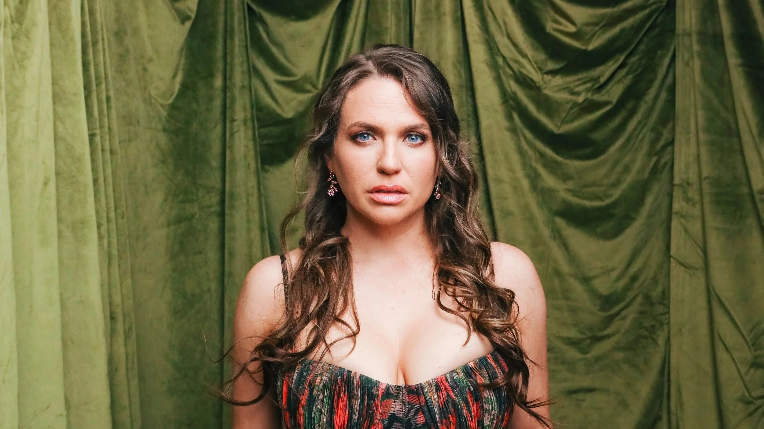 A woman with long, curly brown hair and blue eyes standing in front of green curtains, wearing a colorful strapless dress and pink earrings.