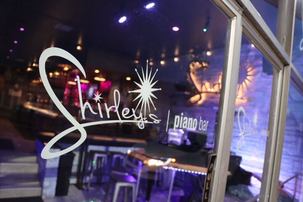 Glass window with the logo for girley's piano bar, featuring a stylized star and the text "girley's" and "piano bar," inside a dimly lit venue with purple and blue lighting.