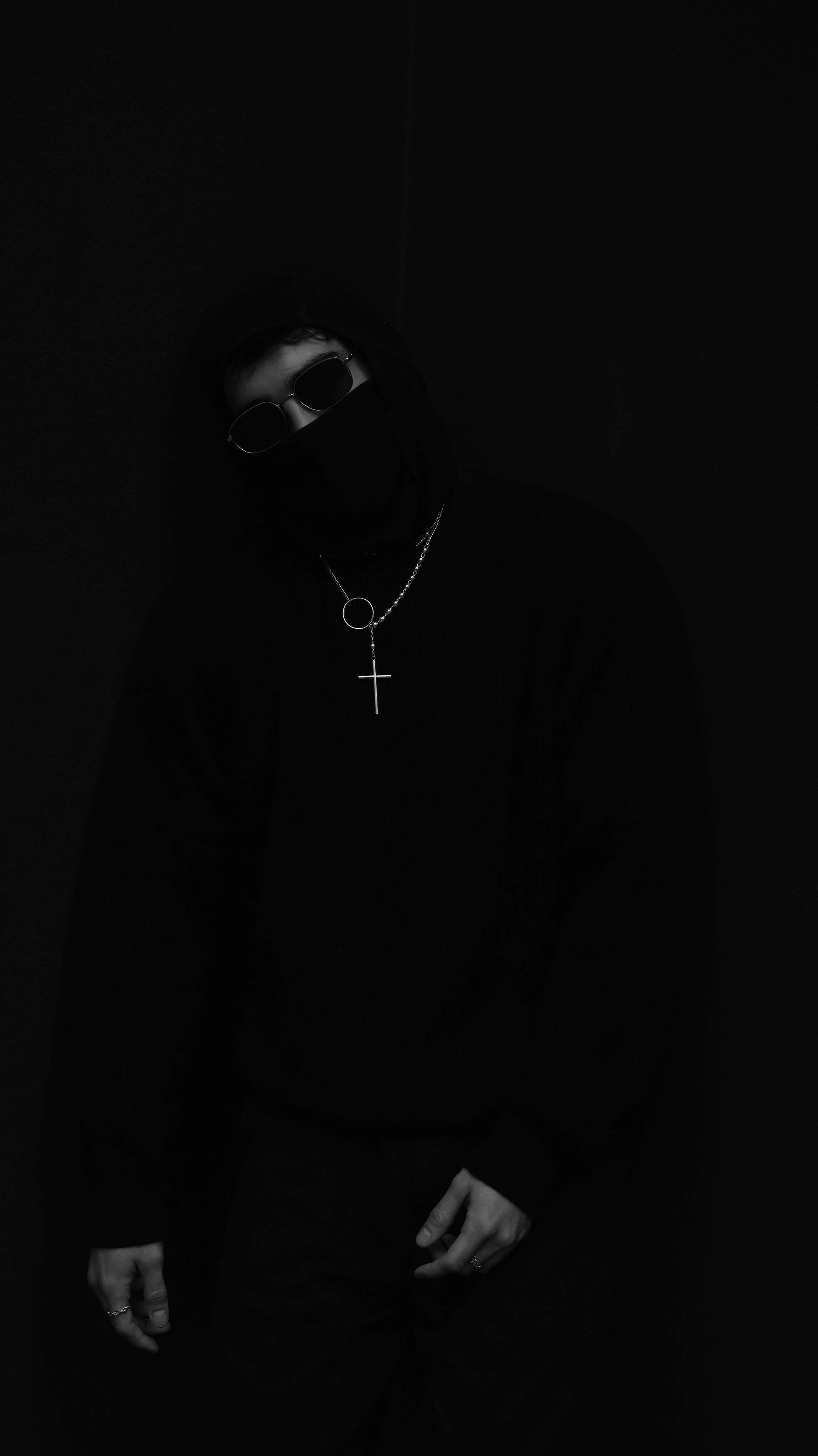 Person wearing dark clothing, sunglasses, and a mask, with necklaces including a cross, standing against a dark background.