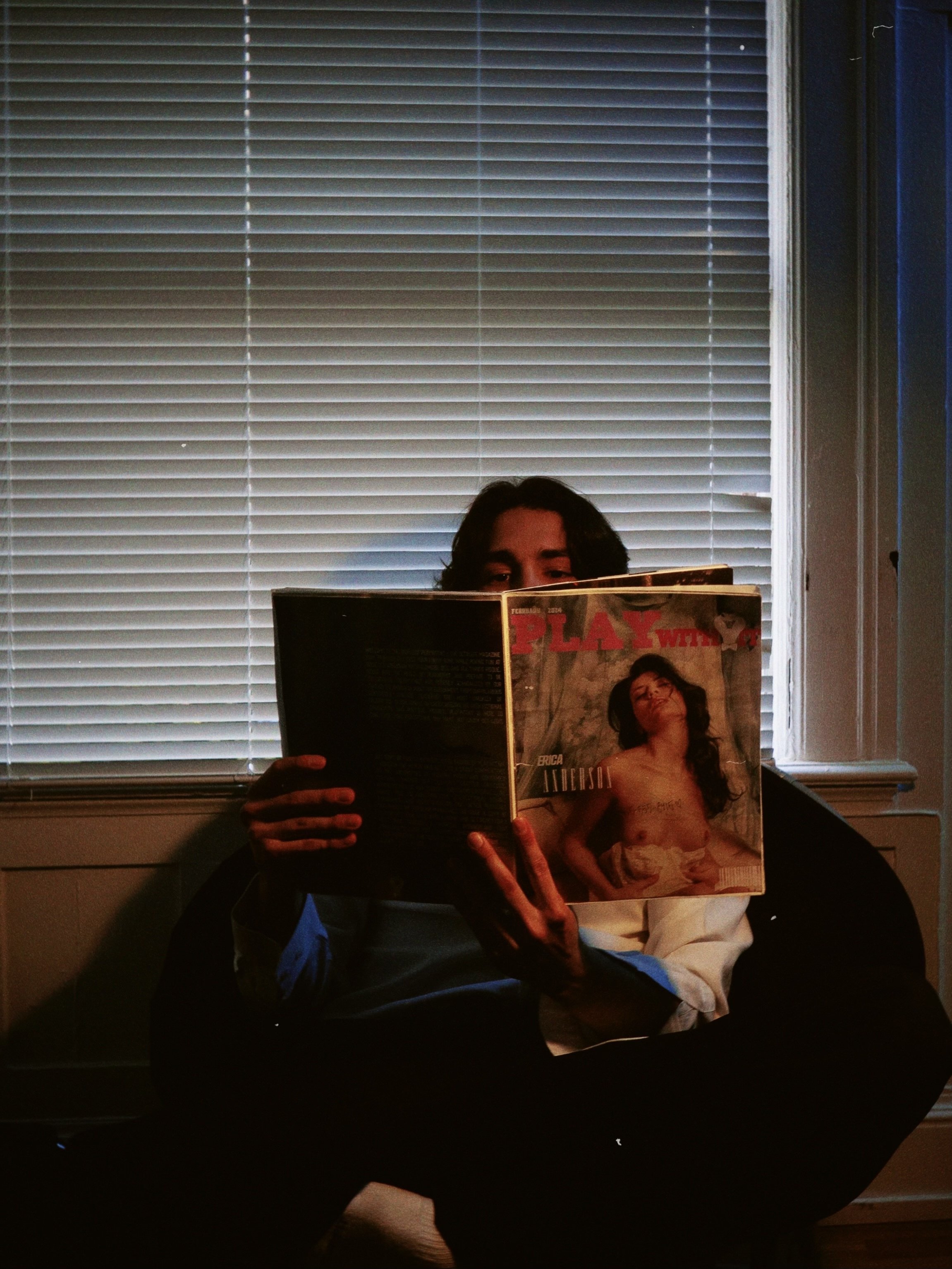 Person sitting on a chair in front of window blinds, reading a magazine with a woman on the cover.