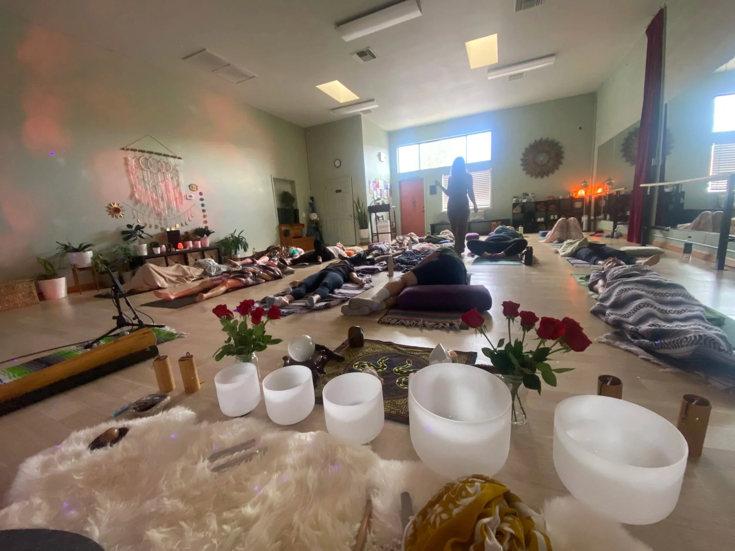 People participating in a yoga or meditation class in a spacious room with soft natural lighting, mats and bolsters on the floor, along with decorative plants, candles, and wall hangings.