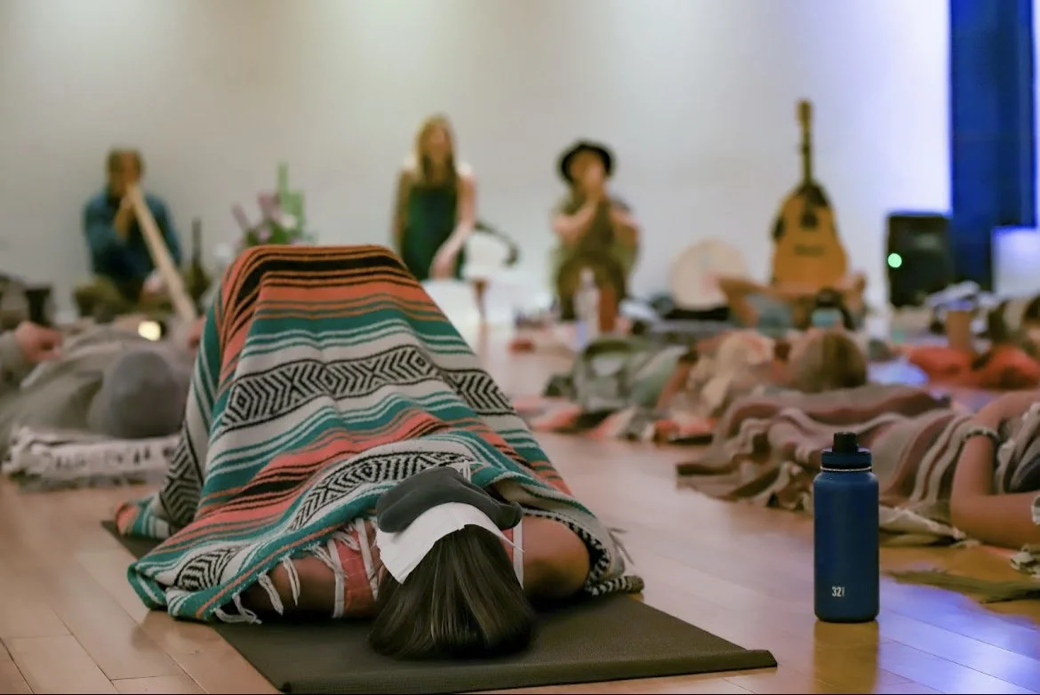 A woman in a blanket meditating on a yoga mat during a group meditation or yoga class, with other participants, a guitar, and musicians in the background.