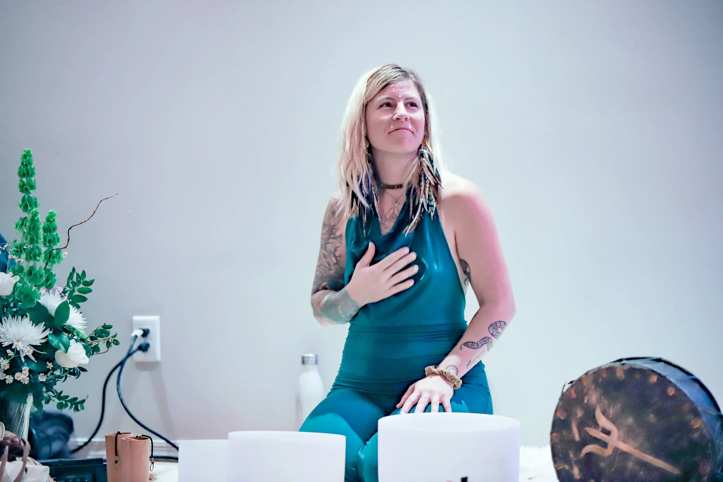 A woman with blonde hair, tattoos on her arms, wearing a teal dress, sitting on a white surface, with her hand on her chest, and looking upwards.