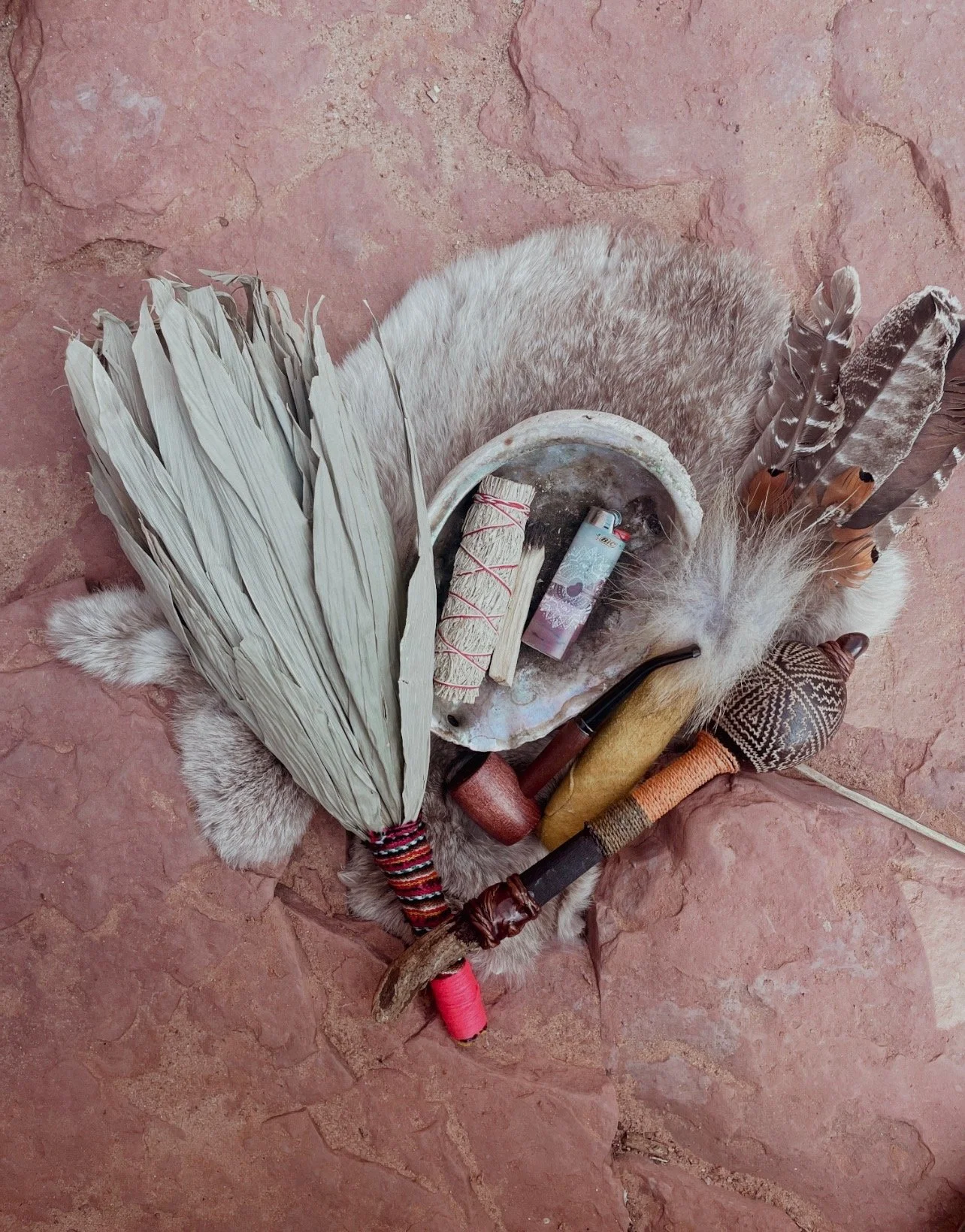 A collection of traditional African ritual items on a red stone floor, including feathers, a pipe, a small drum, and decorated sticks.