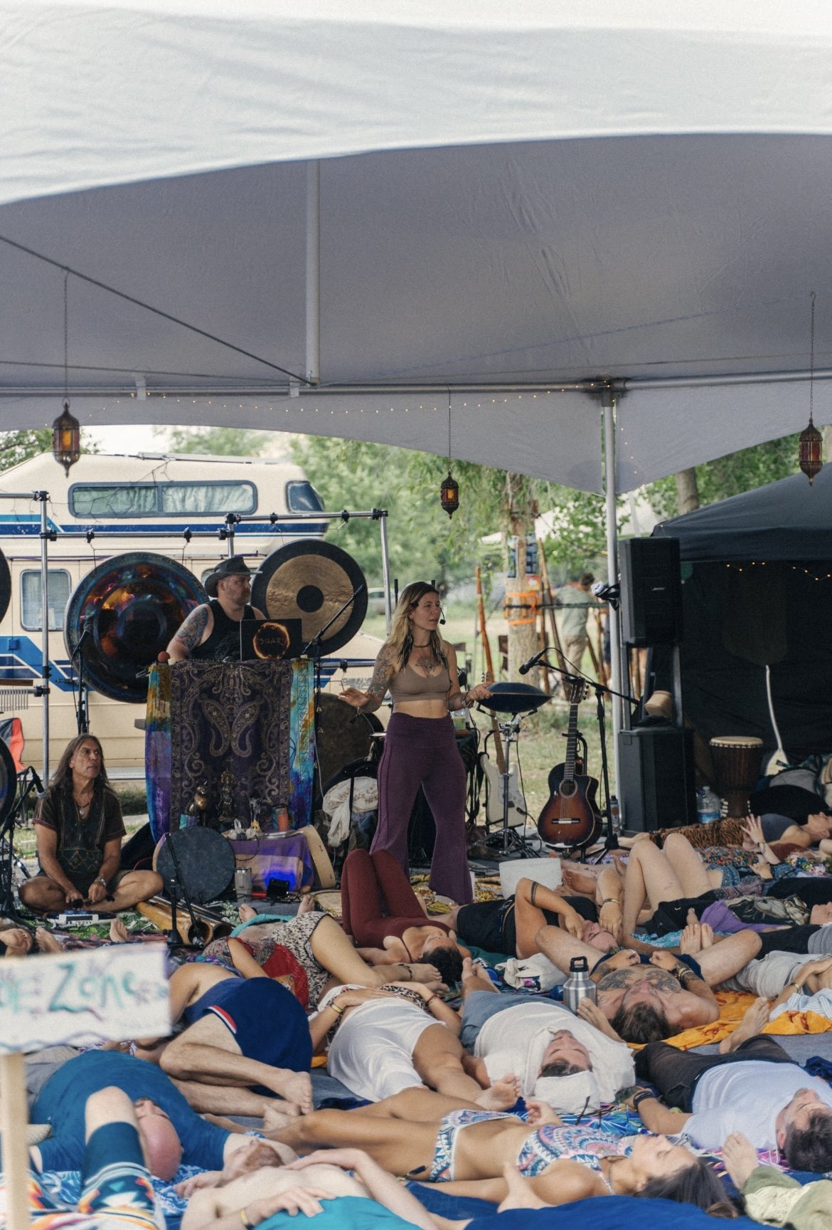 People attending an outdoor event or festival, lying on mats and blankets, with a band performing on a tented stage in the background.