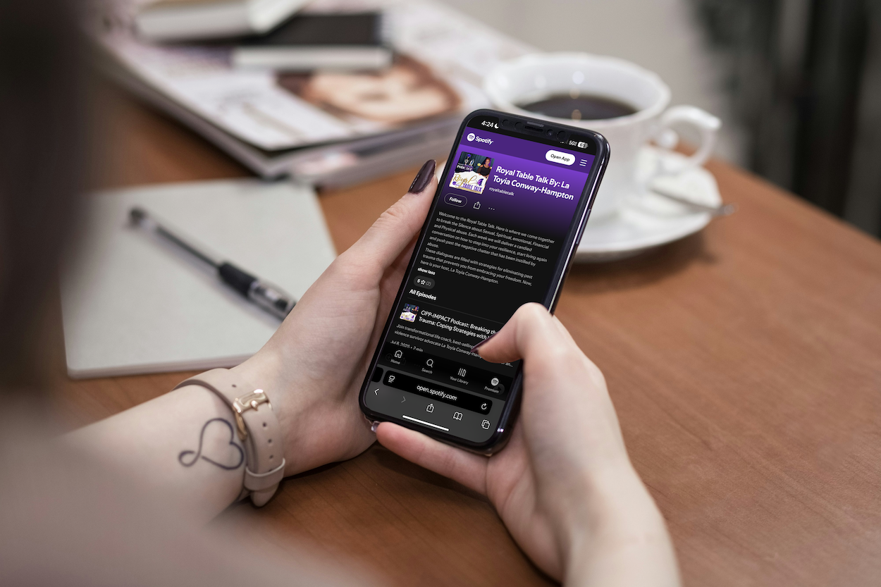 Person holding an iPhone with Spotify app open, displaying a podcast titled 'Royal Table Talk by La Toya Conway-Hampton'; a notebook, pen, magazine, and a cup of coffee on a wooden table.
