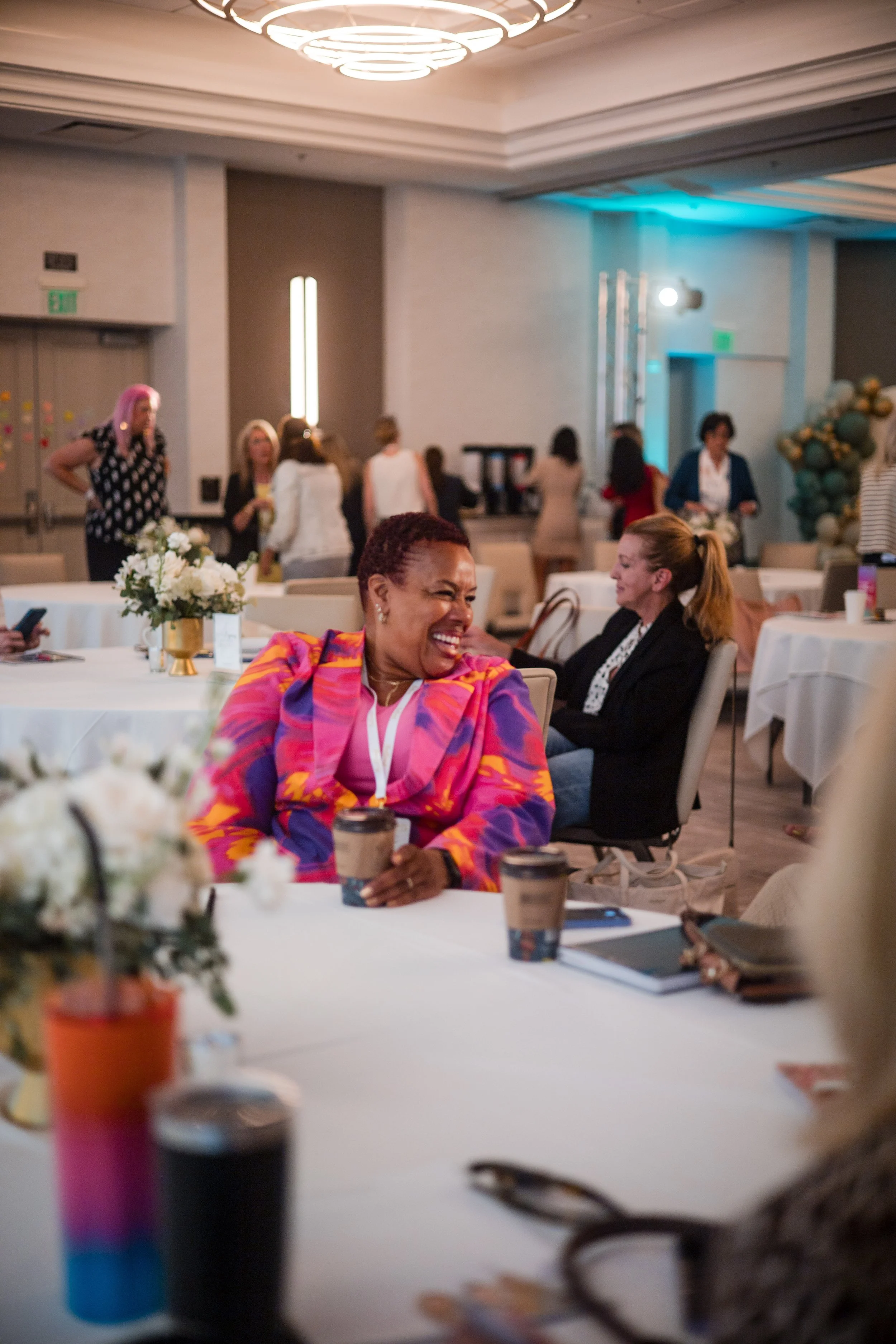 A group of women at a social event, with one woman in a colorful pink and purple patterned blazer laughing at a round table decorated with flowers, coffee cups, and notebooks.