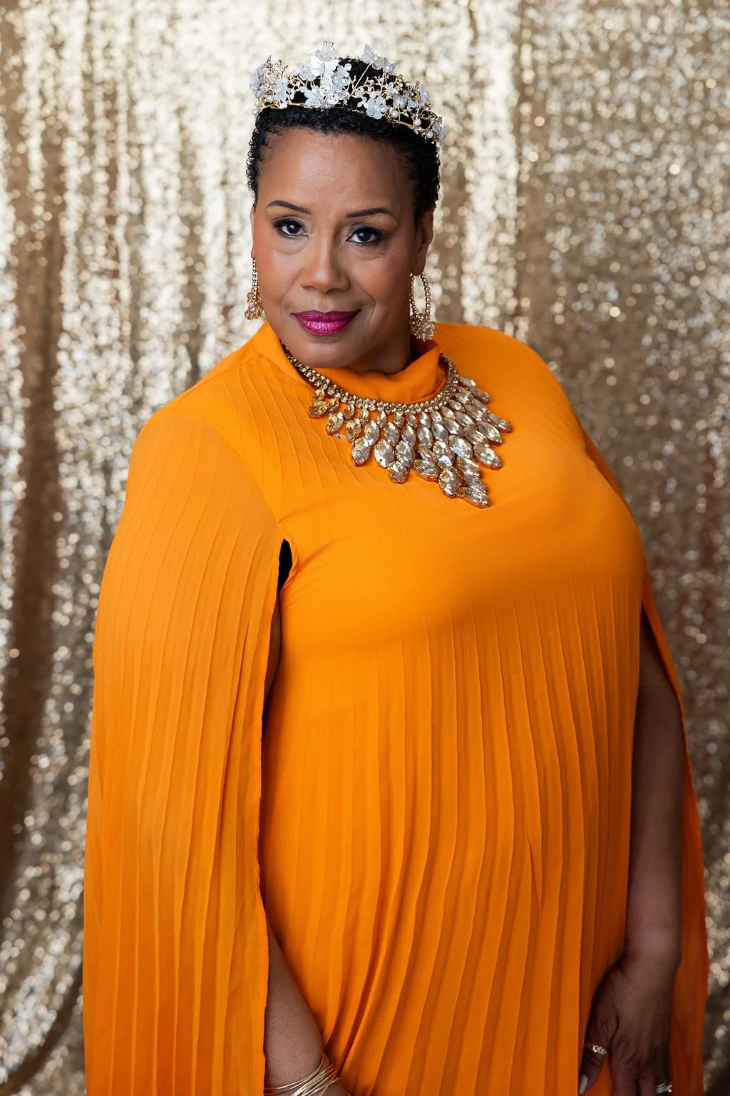 A woman wearing an orange pleated dress, a sparkling tiara, and a large diamond necklace and earrings, standing against a gold sequin backdrop.