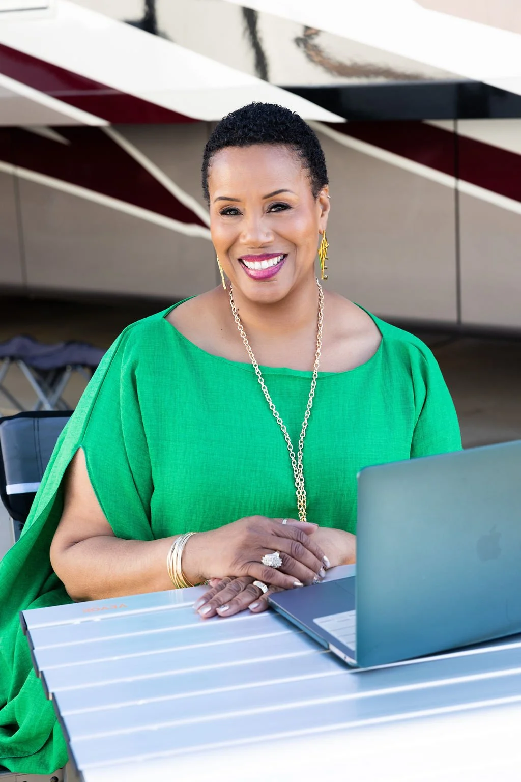 Woman with short curly hair wearing a green dress, sitting at a table with a laptop, smiling, outdoor setting.