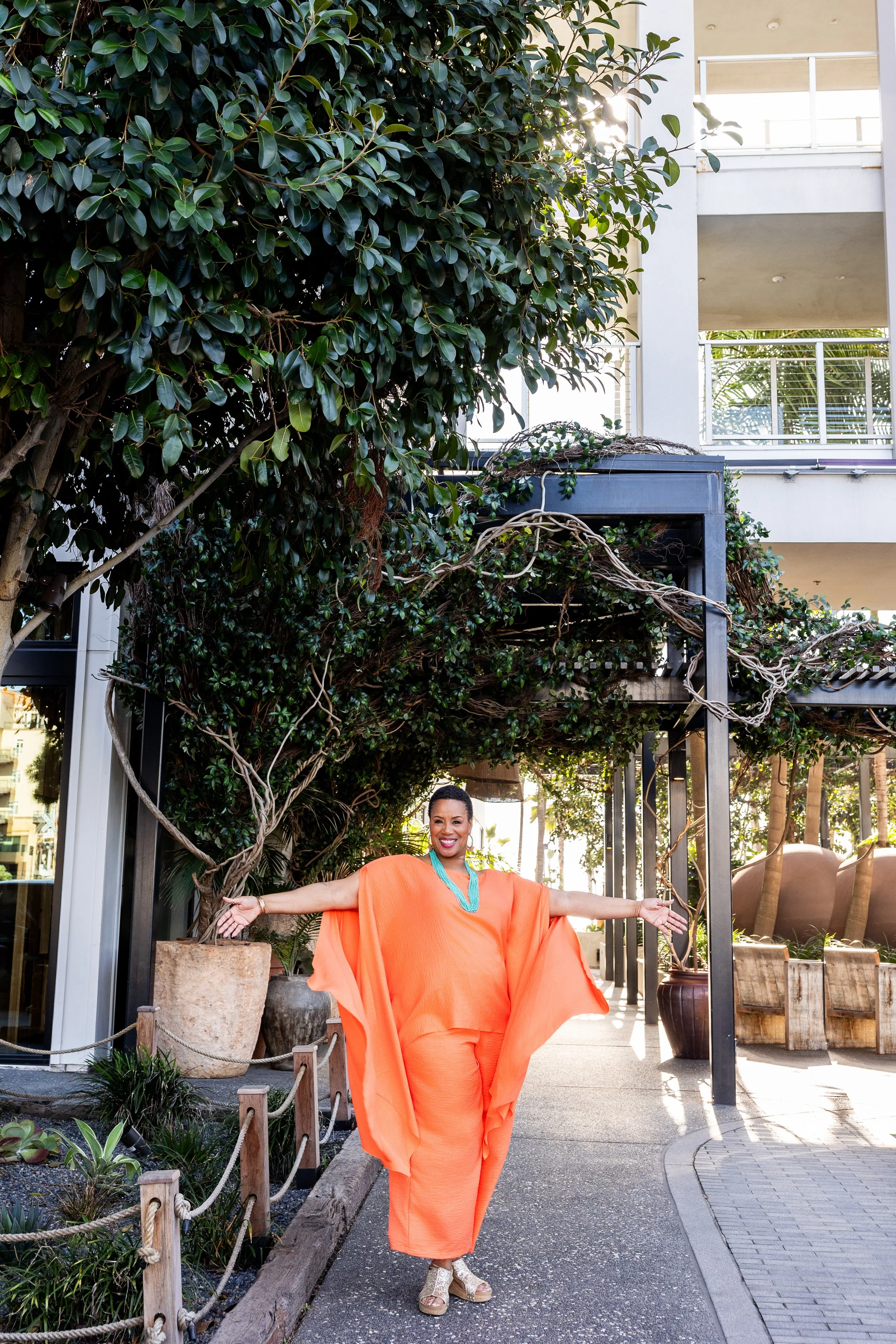 A woman dressed in orange with arms outstretched, standing under a trellis with intertwined vines and green leaves, smiling outdoors.