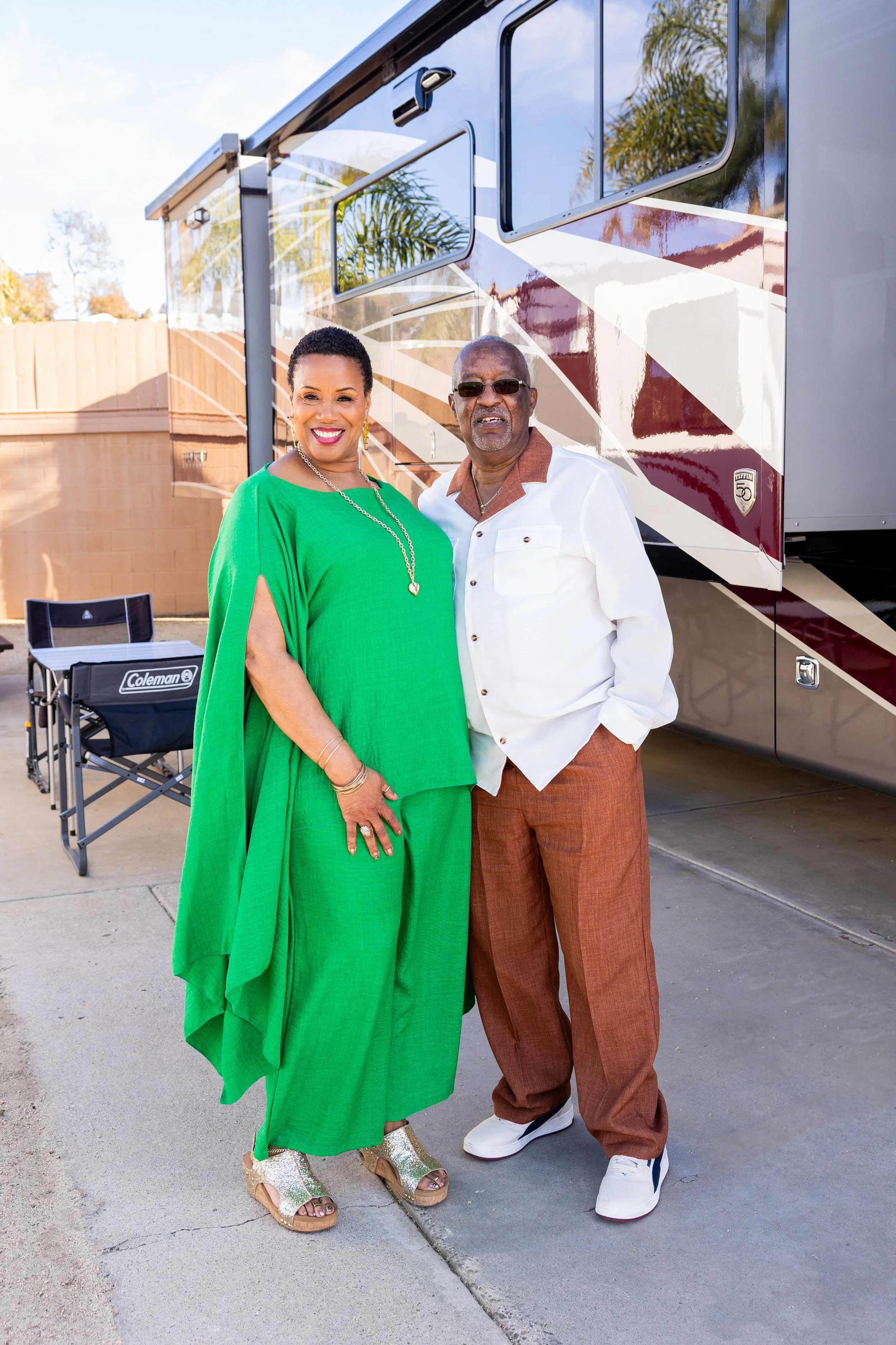 A woman in a bright green outfit with gold jewelry and sandals stands next to a man wearing sunglasses, a white shirt with brown collar, brown pants, and white sneakers, standing outdoors in front of a large RV.