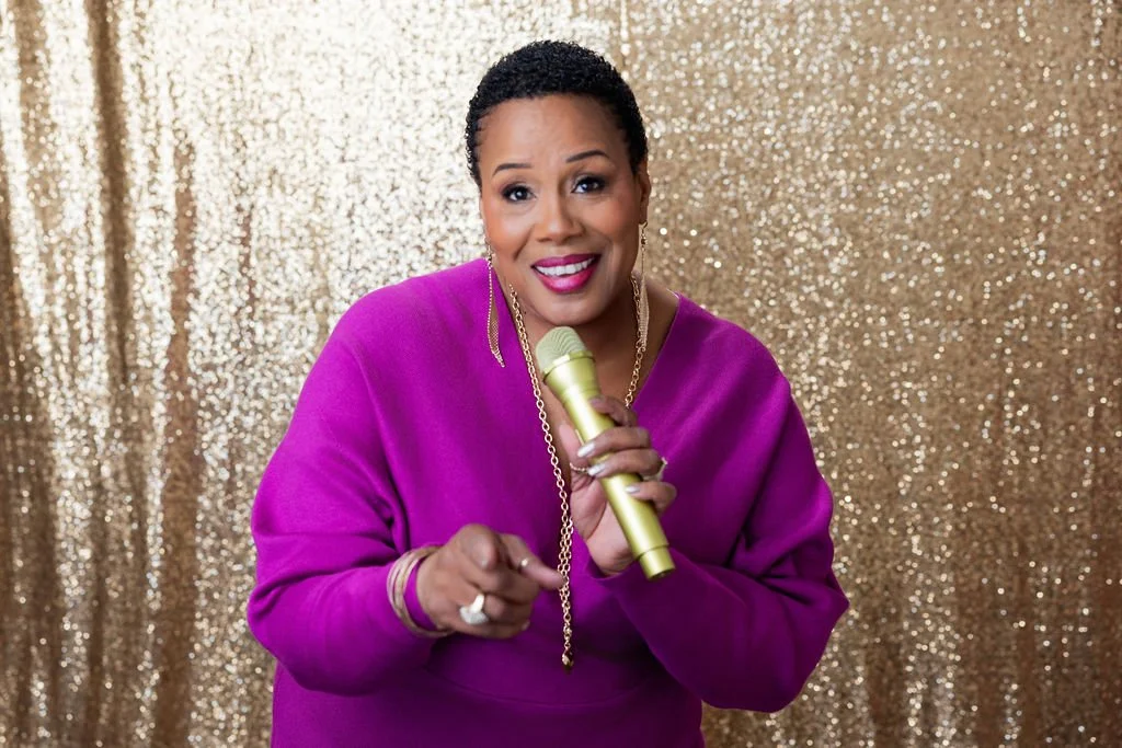 A woman with short black hair wearing a purple sweater, holding a gold microphone, pointing with her finger, standing in front of a gold glitter background.
