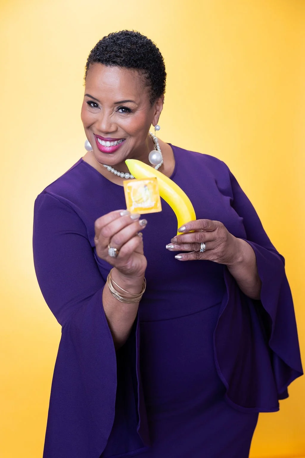 A smiling woman holding a banana, a small orange drink, and a lemon-shaped object against a yellow background.