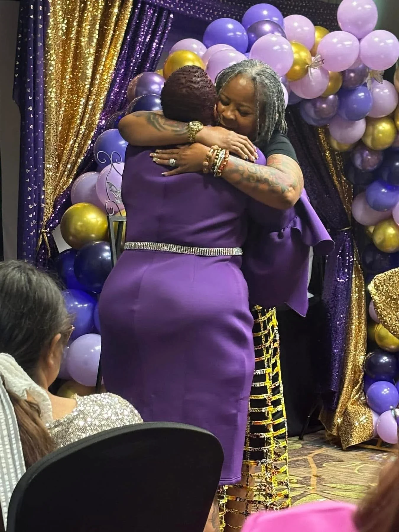 Two women hugging at a celebration with purple, gold, and black balloons, and decorative backdrop.