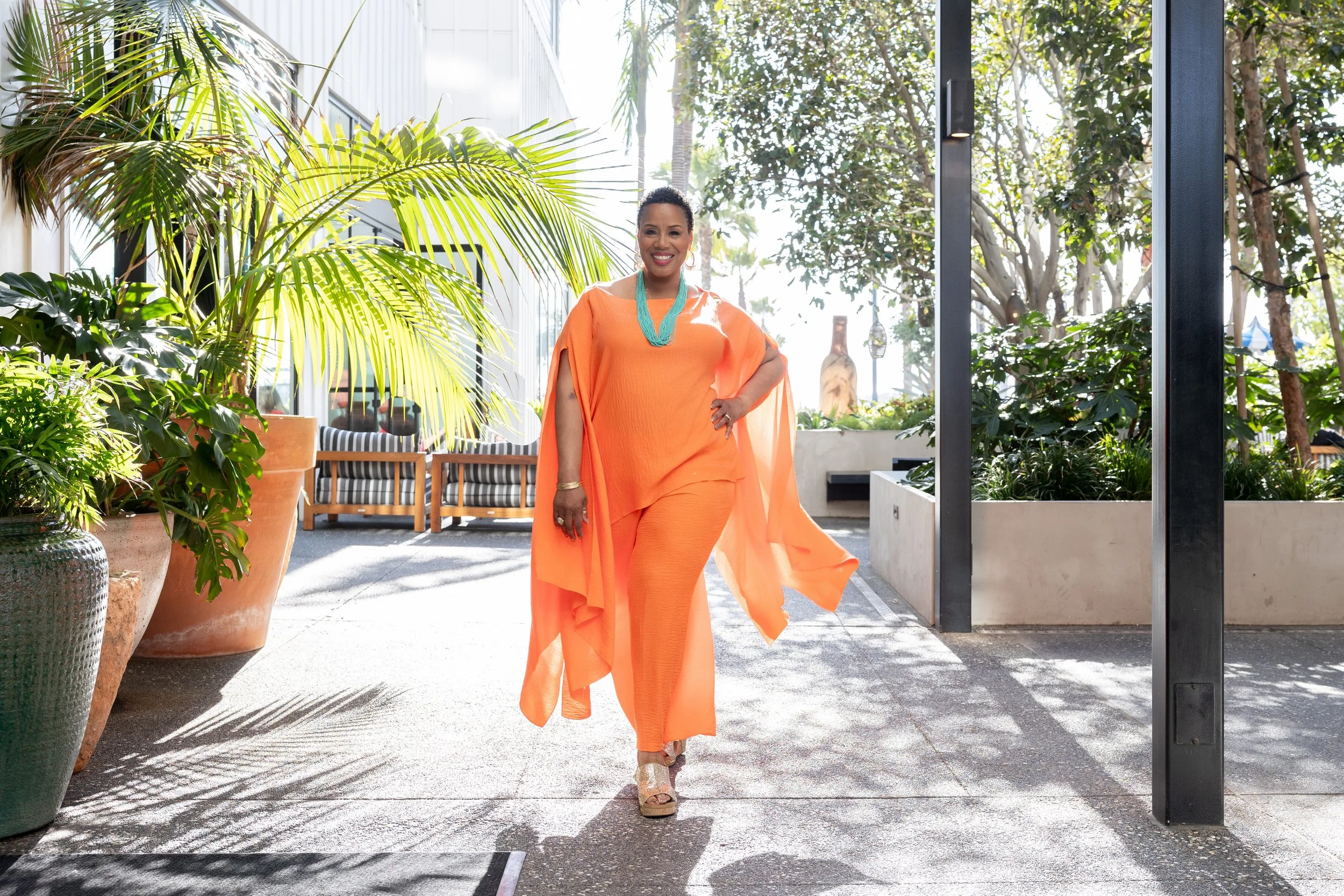 Woman in orange outfit walking outdoors among potted plants and trees, smiling and wearing turquoise necklace and platform sandals.