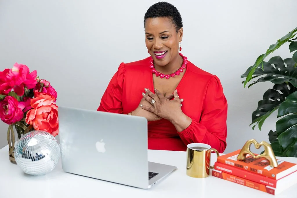 A woman in a red dress with jewelry sitting at a white desk, looking at a laptop with a smile, surrounded by flowers, books, a gold mug, and a gold heart sculpture.