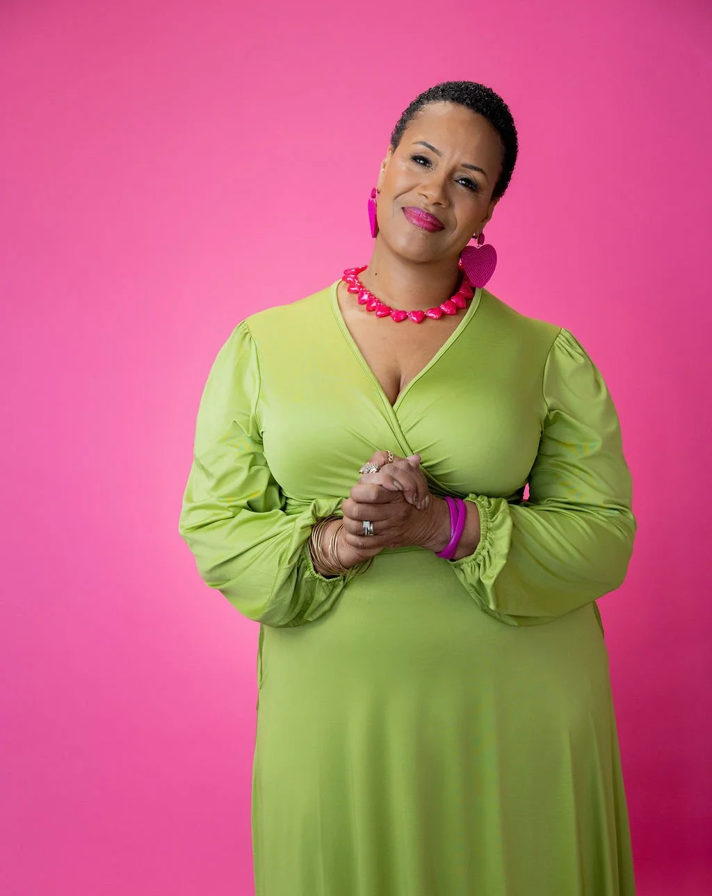 A woman with short curly hair, dressed in a green dress, standing against a pink background. She is wearing pink heart-shaped earrings, a pink necklace, and pink bracelets, and has a confident and friendly expression.