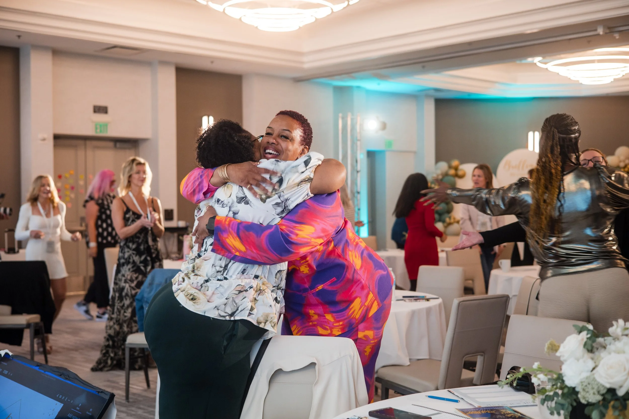 Two women hugging and smiling at a celebration or event, with other guests in the background celebrating.
