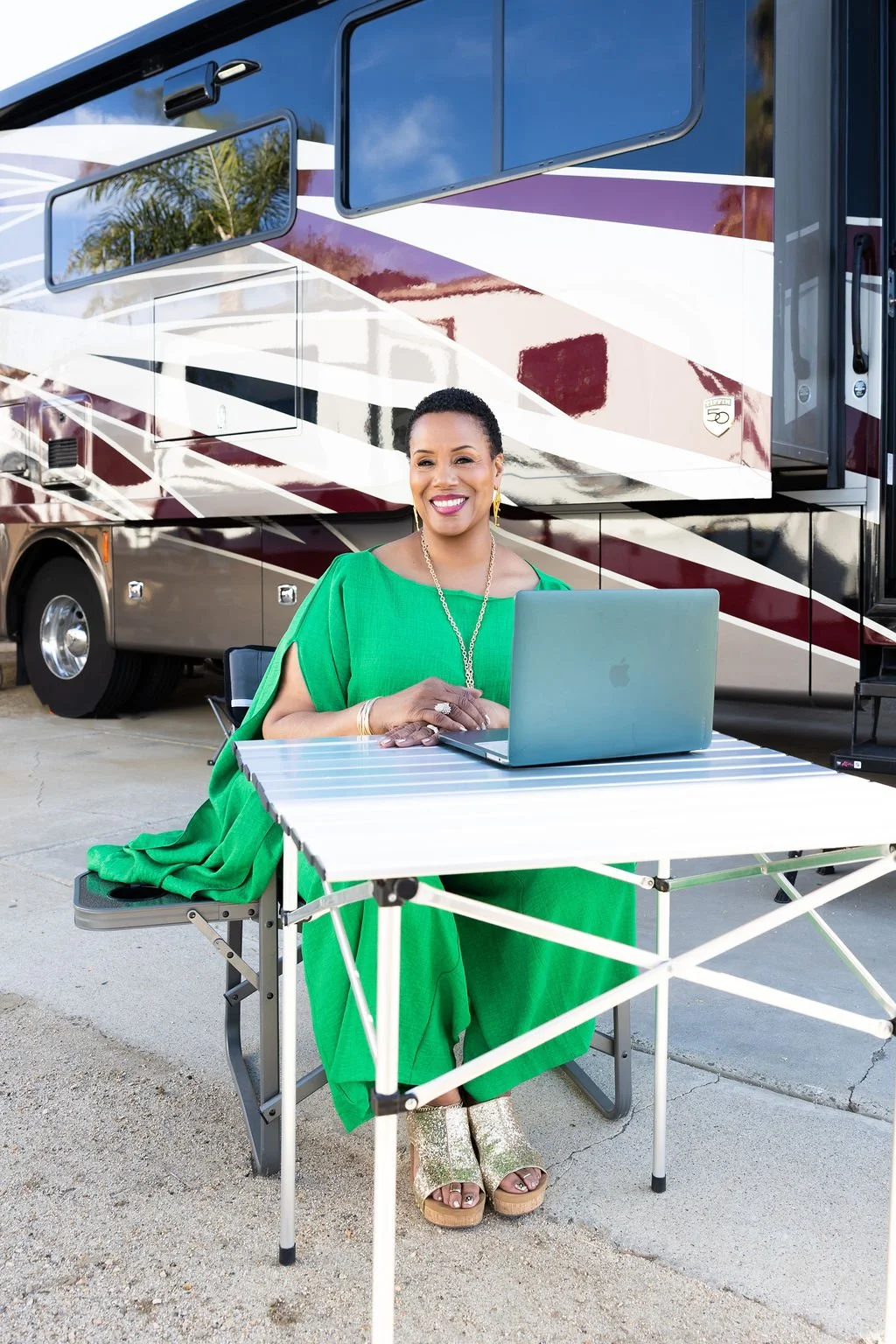 A smiling woman in a bright green dress working on a silver laptop, seated at a portable table outside a large RV.