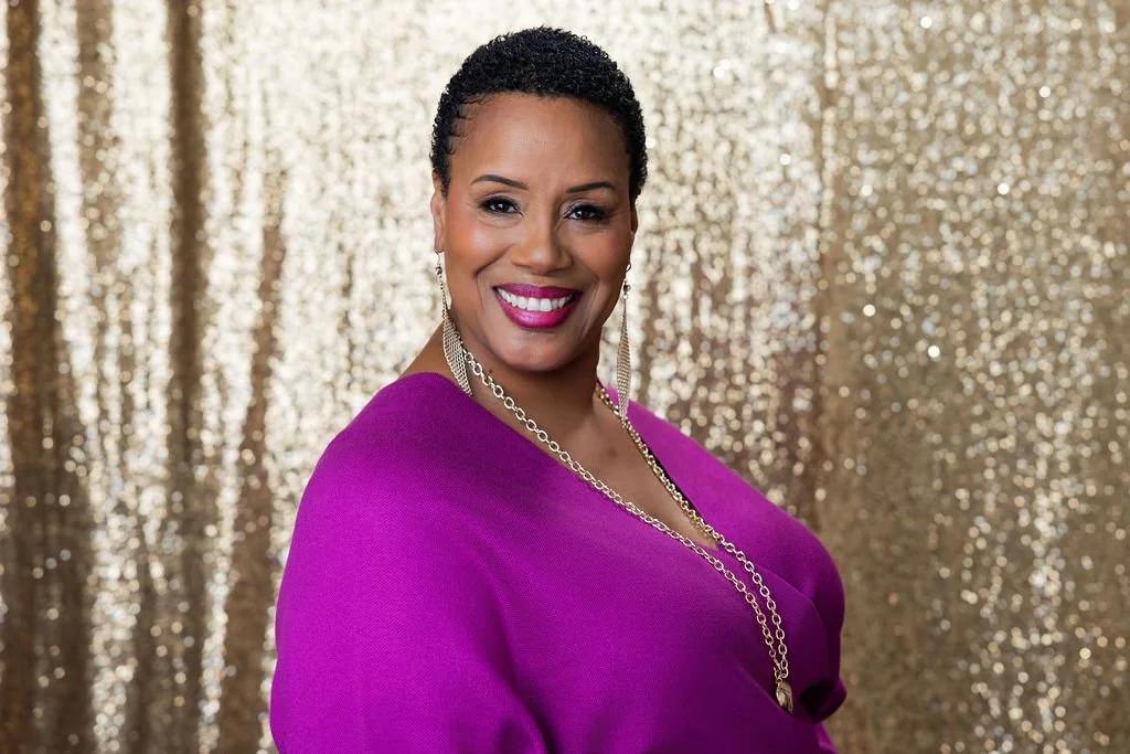 A smiling woman with short, curly dark hair, wearing a purple top, gold jewelry, and standing in front of a gold, glittery background.