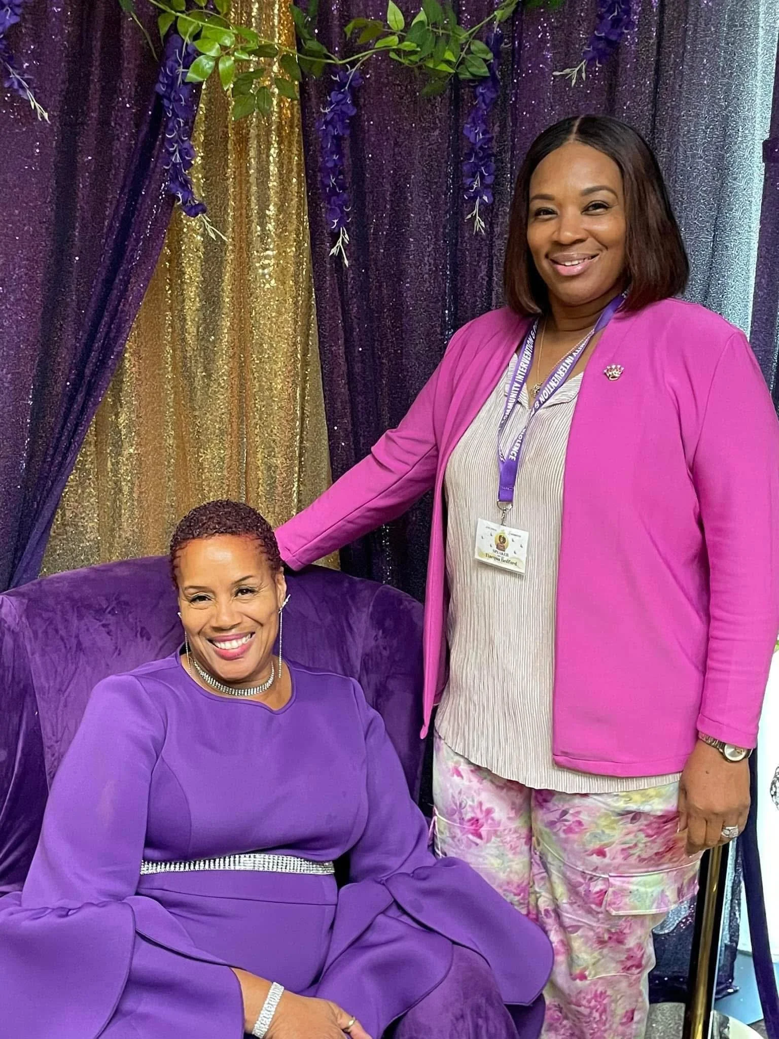 Two women, one seated and one standing, in front of purple, gold, and sparkly curtains decorated with purple flowers and green leaves. The seated woman is wearing a purple dress with a rhinestone belt, and the standing woman is dressed in a pink blazer with a lanyard around her neck and floral pants. Both women are smiling.