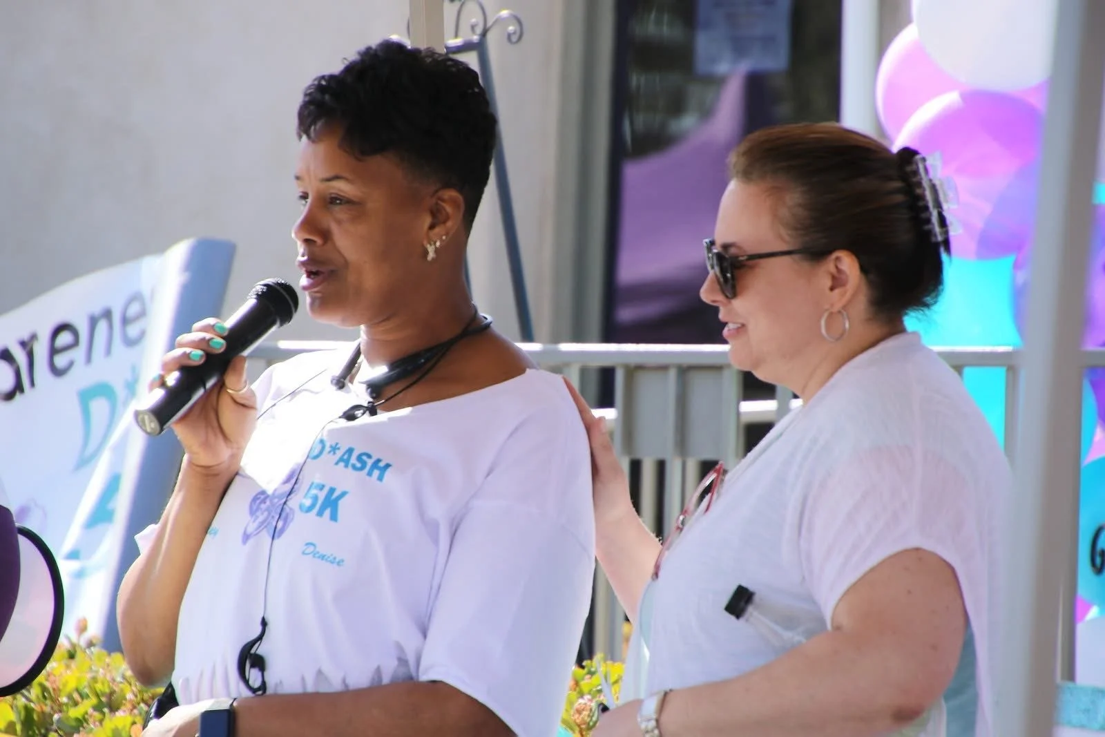 Two women at an outdoor event, one speaking into a microphone, the other gently touching her shoulder. One woman has short dark hair and is wearing a white shirt with blue text, the other has brown hair in a bun, sunglasses, and a white shirt. There are purple and white balloons in the background.