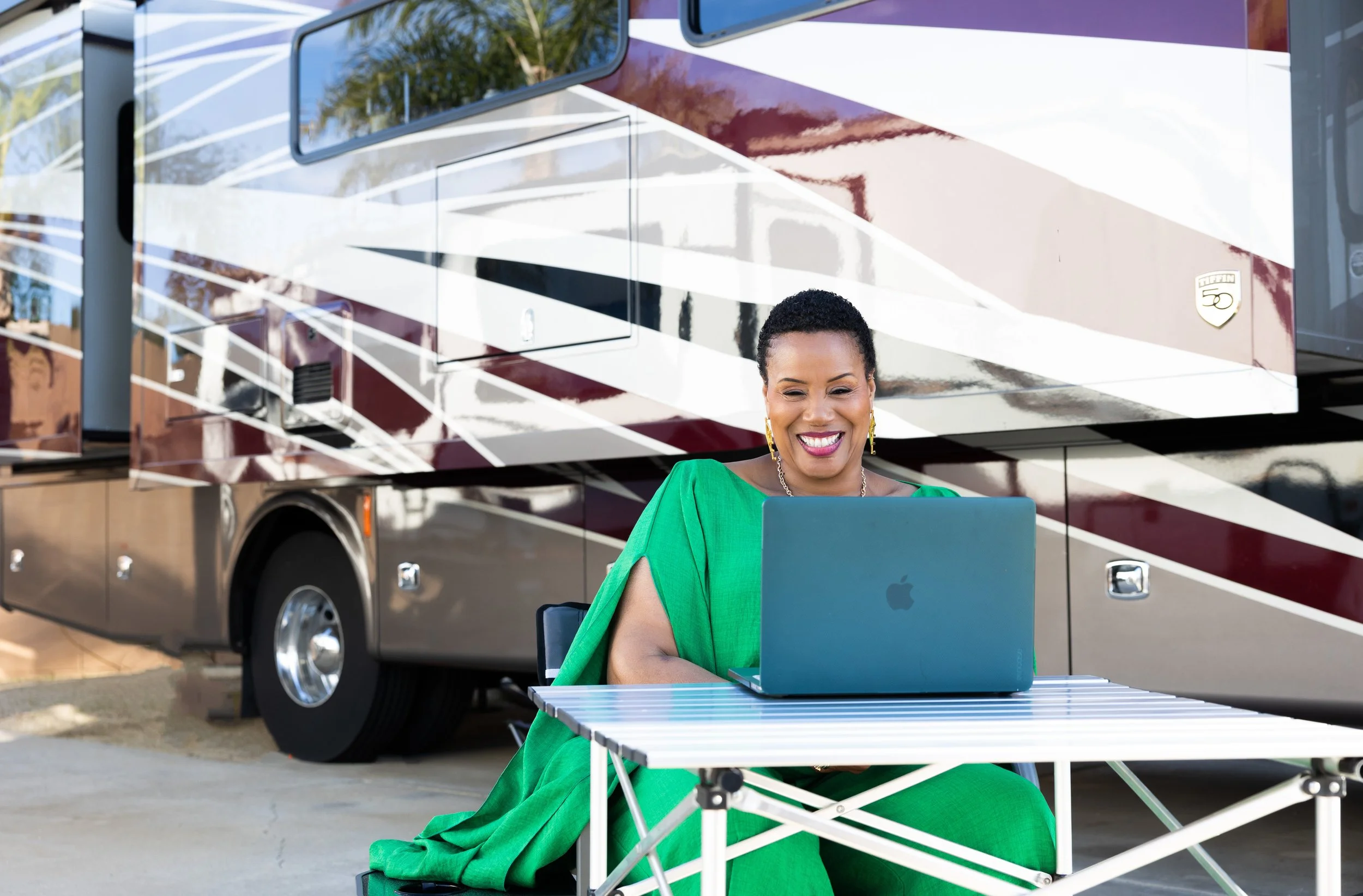 A woman sitting at a table outdoors, using a MacBook, in front of a luxury RV with a shiny exterior and distinctive design.