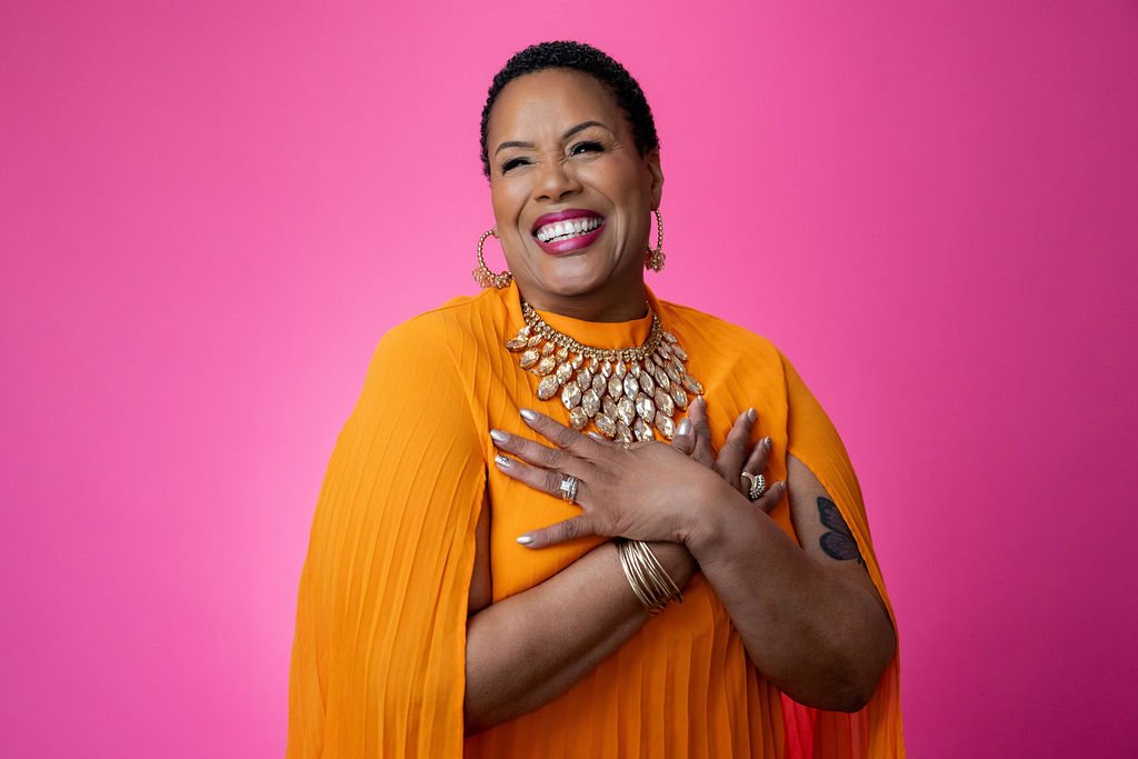 A joyful woman wearing an orange dress and large jewelry, smiling with hands crossed over her chest, against a pink background.