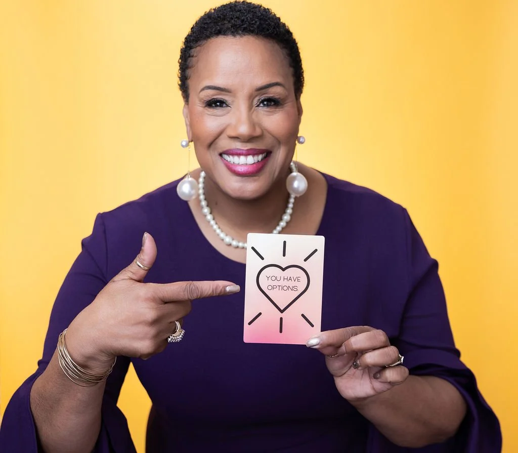 A woman with short curly hair, wearing a purple top, pearl earrings, a pearl necklace, and jewelry, is smiling and pointing to a card that says 'You have options' with a heart design on it, against a yellow background.
