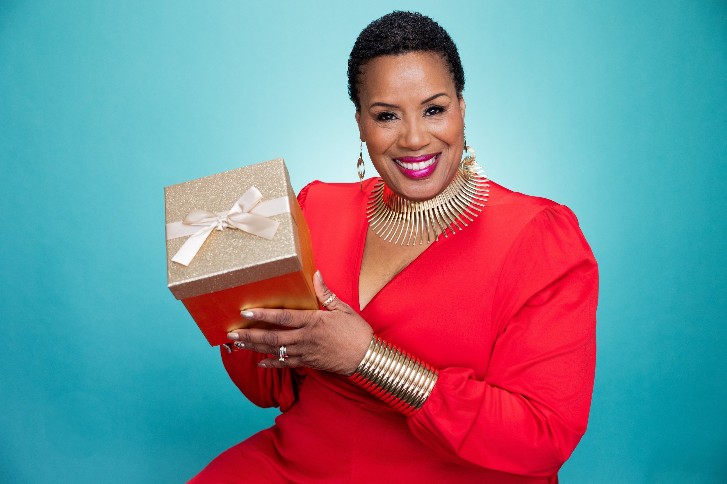 A smiling woman with short curly hair wearing a red dress and gold jewelry holding a gift box with a gold glitter top, white ribbon, and bow in front of a blue background.