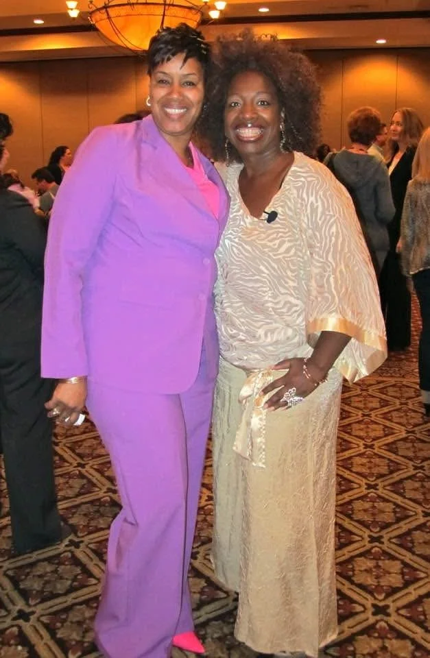 Two women smiling and posing together at an indoor event. One woman is wearing a purple suit, and the other is wearing a cream-colored top with a matching skirt. There are other people in the background.