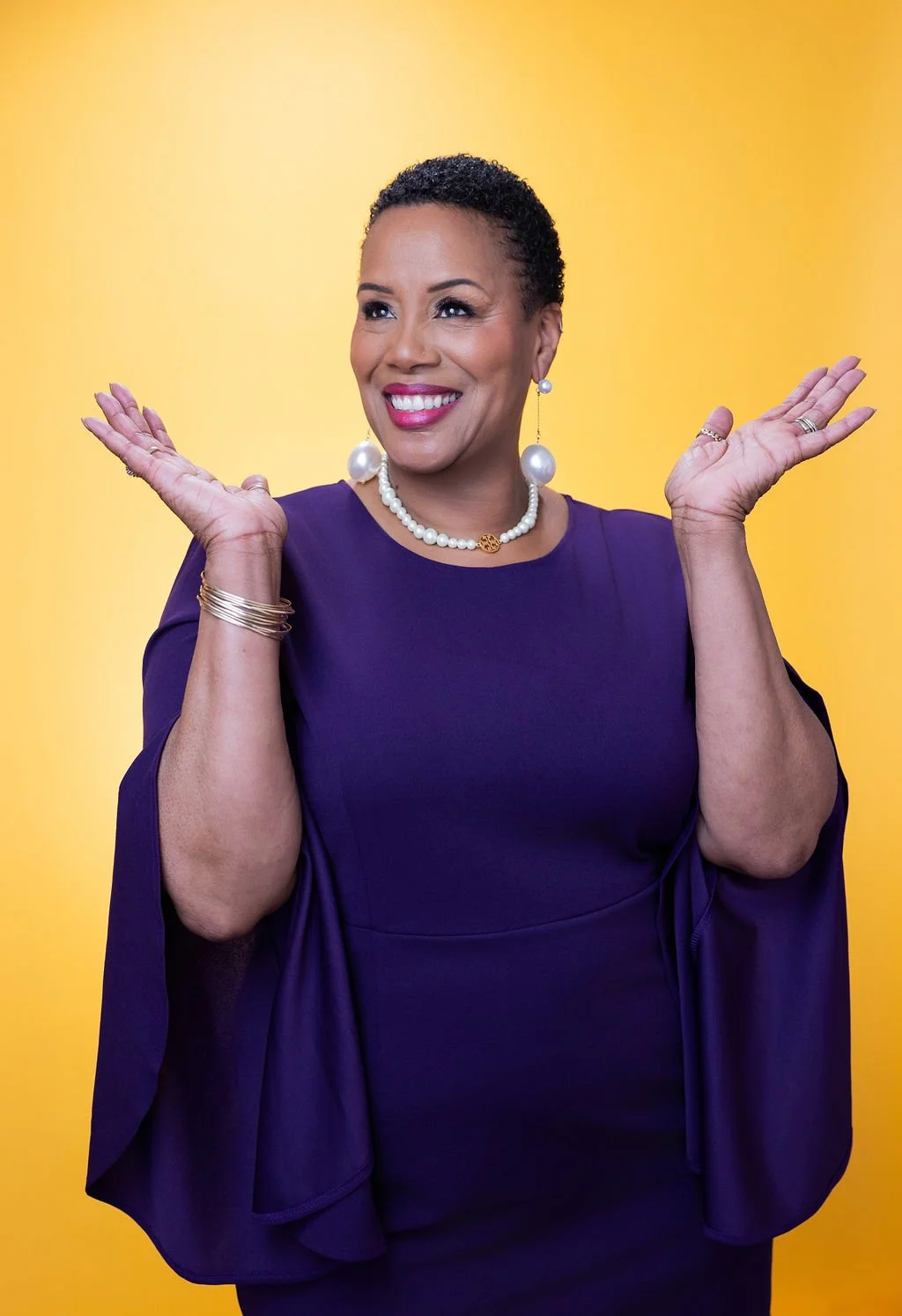 A smiling woman with short curly hair wearing a purple dress, pearl necklace, pearl earrings, and gold jewelry, posing against a yellow background.