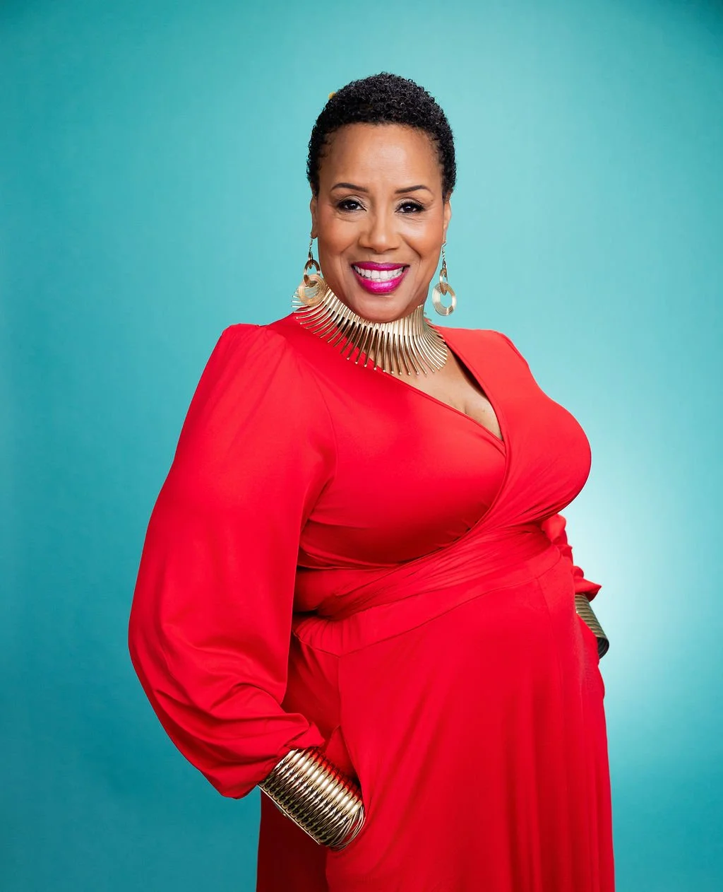 A woman with short curly hair wearing a red dress, large gold jewelry including a neck piece, earrings, and bracelets, standing against a teal background with a smile.