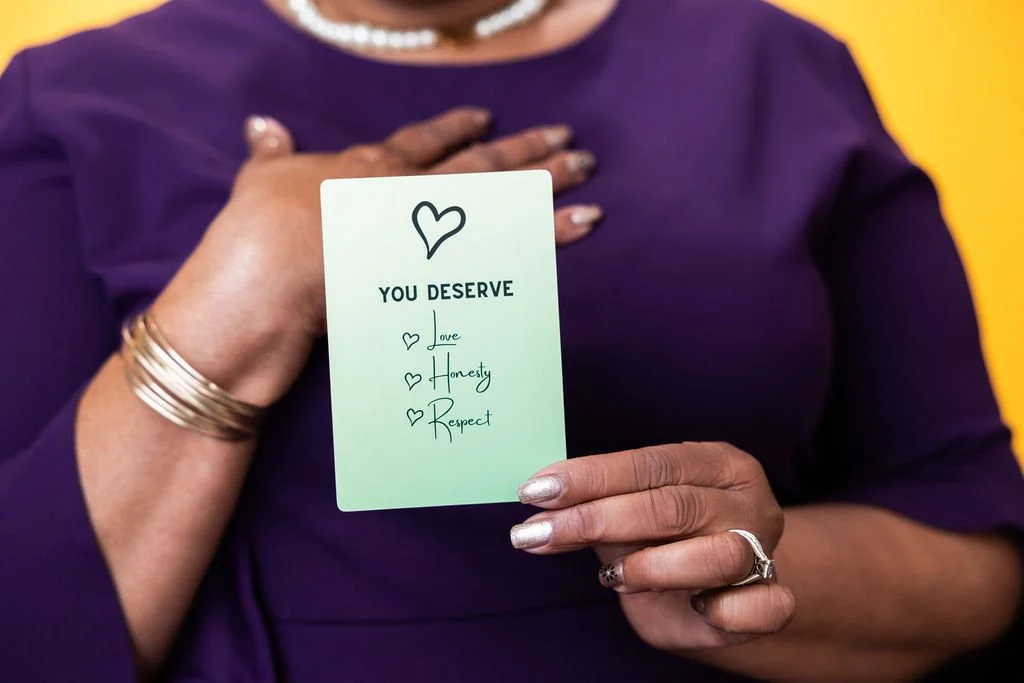 A person holding a card that reads 'You Deserve love, honesty, respect' with a hand placed on their chest. The person is wearing a purple outfit, jewelry, and has painted nails, against a yellow background.