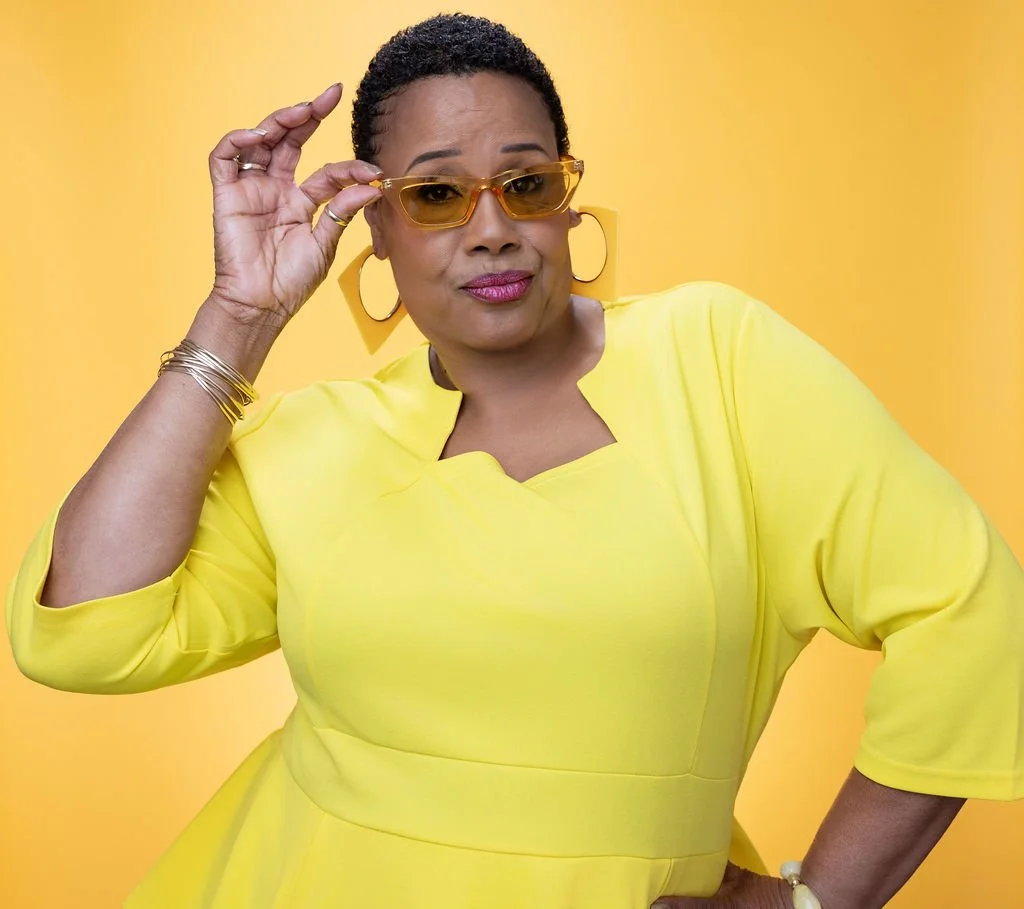 A woman with short curly hair wearing a yellow dress, yellow sunglasses, large hoop earrings, and gold jewelry, striking a confident pose against a yellow background.
