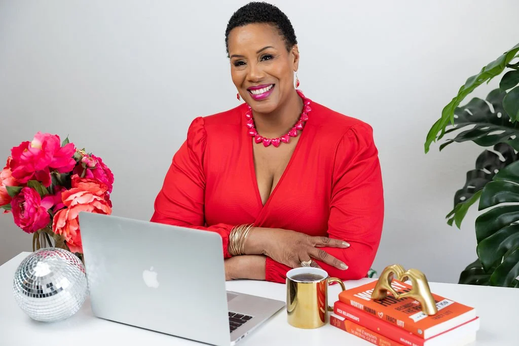 A smiling woman in a red dress sitting at a desk with a laptop, pink flowers, a gold cup of coffee, a disco ball, and orange books, with green plants in the background.