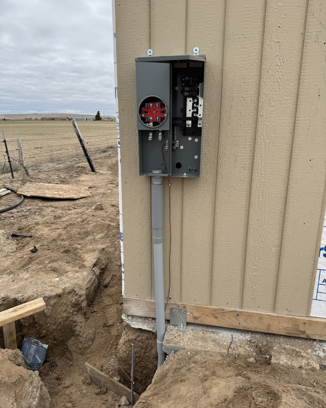 An outdoor electrical meter box mounted on the side of a house, with exposed wiring and conduit, in a rural area with open fields and cloudy sky in the background.