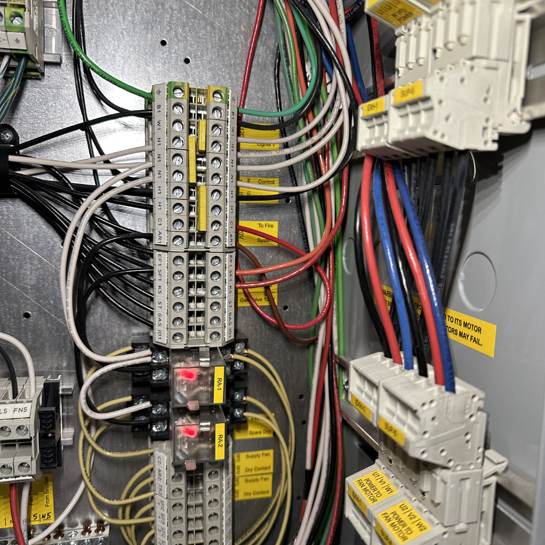 Close-up of an electrical control panel with various wires, terminal blocks, and relays, labeled with yellow tags for different functions.