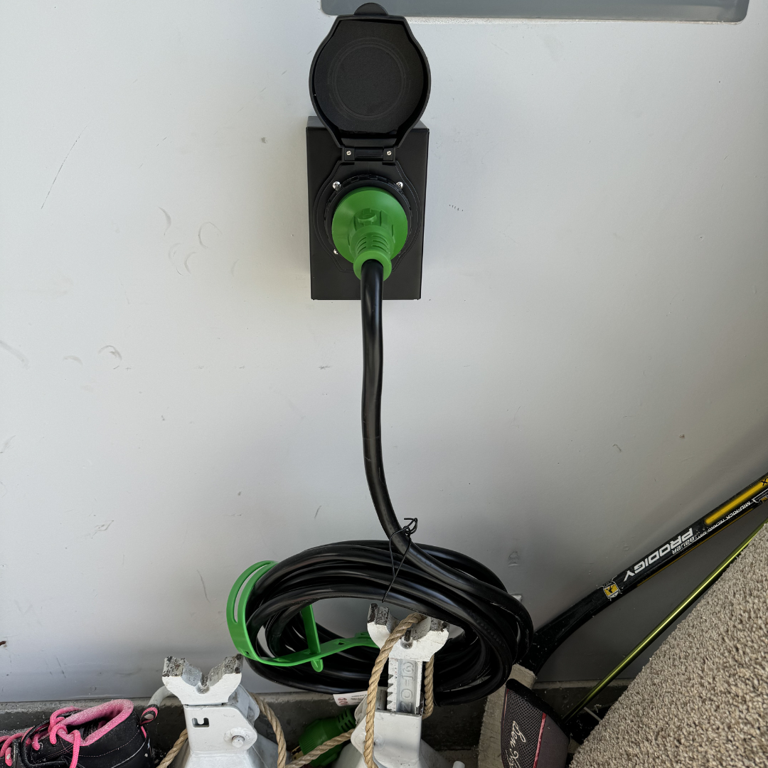 An electrical outlet with a green electric vehicle charging plug inserted, connected to a coiled black cable, and a yellow and black hockey stick resting against the wall.