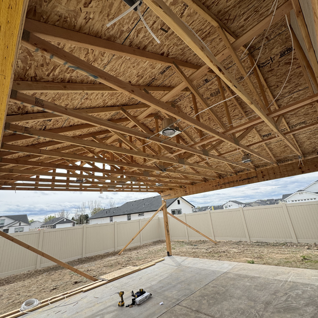 Construction site with a partially built wooden roof structure, exposed beams and plywood sheathing, overlooking a backyard with a concrete floor and a beige privacy fence, houses visible in the background under a partly cloudy sky.