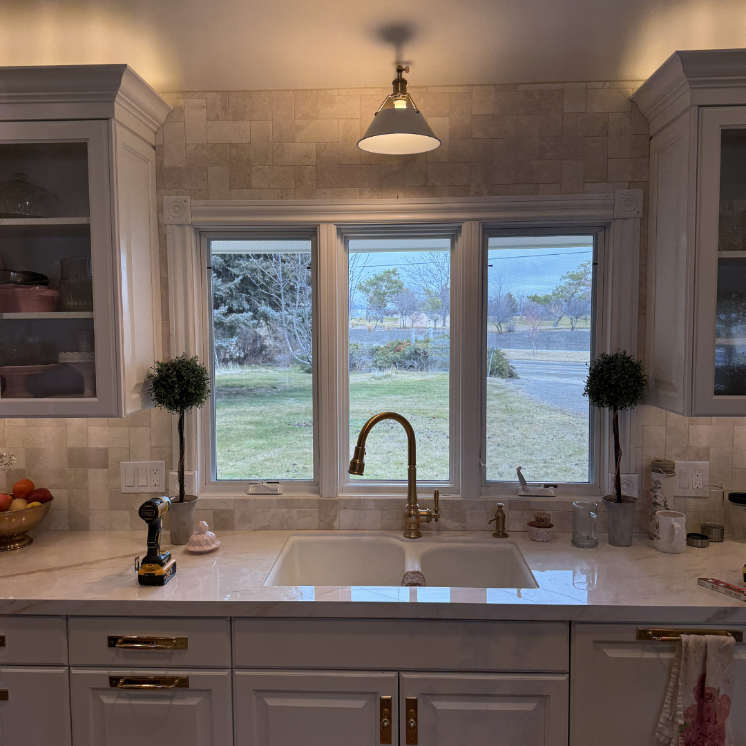 Kitchen window with three panes overlooking a yard with grass, trees, and a water body in the distance. The sink area beneath the window has a brass faucet, small potted topiary plants, and various kitchen items.