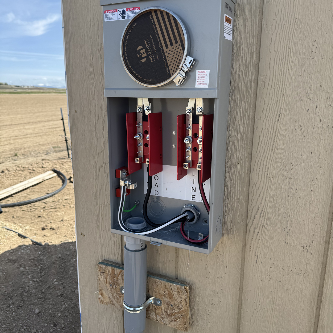 An outdoor electrical box with open cover, revealing wiring and electrical components, attached to a beige wooden wall near a dirt field with a fence in background.