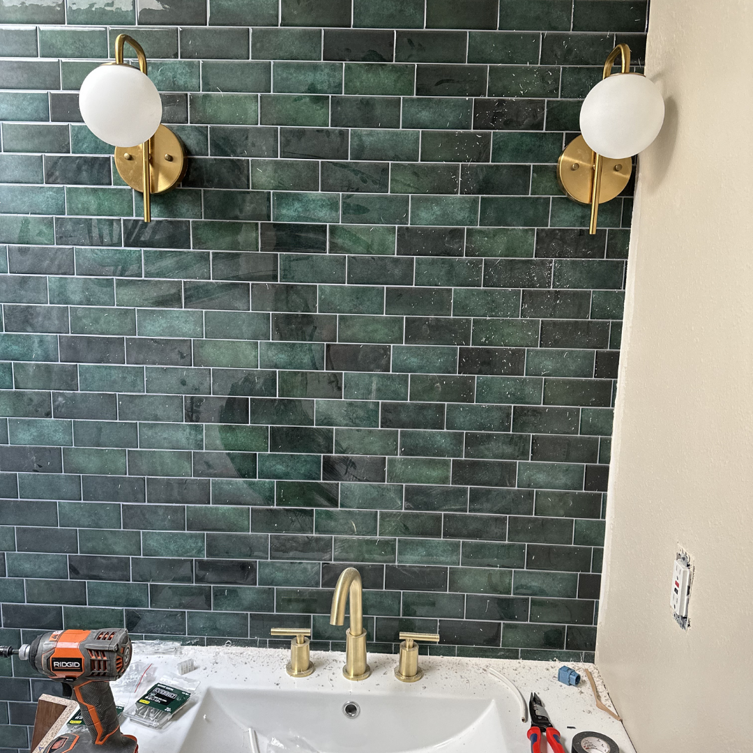 Bathroom with green tiled wall, two brass and white wall sconces, white sink with brass fixtures, and tools including a cordless drill and pliers on the countertop.