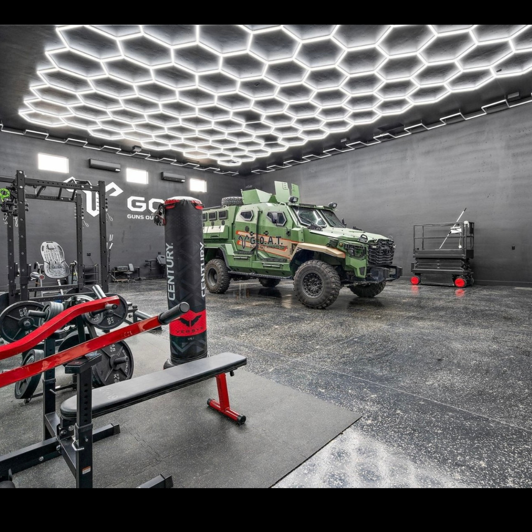 An indoor gym with workout equipment, a punching bag, and a military-style green armored vehicle parked inside under a modern hexagonal LED ceiling.