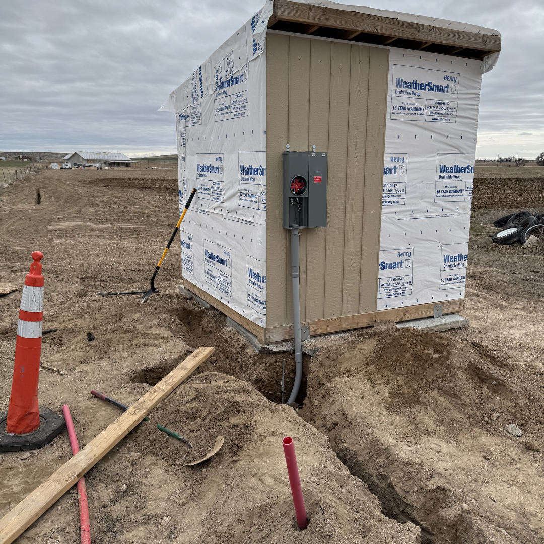 Small construction shed on a dirt site with an electrical box mounted on the outside. Construction tools, pipes, and an orange safety cone are nearby, with a partially buried trench in the foreground. Overcast sky with distant buildings and fields.