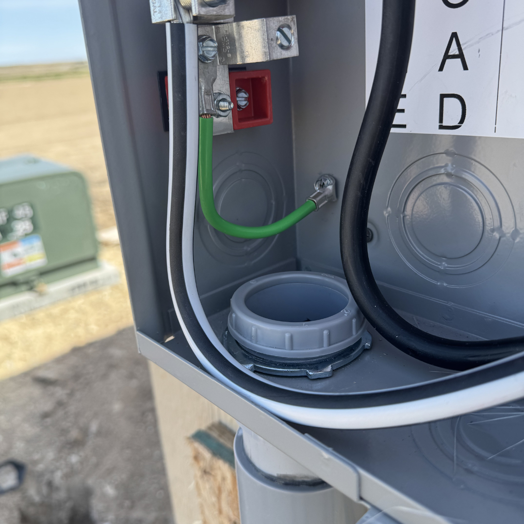 Open electrical box with green and black cables, a gray conduit, and a white threaded cable entry, mounted outside with a field in the background.