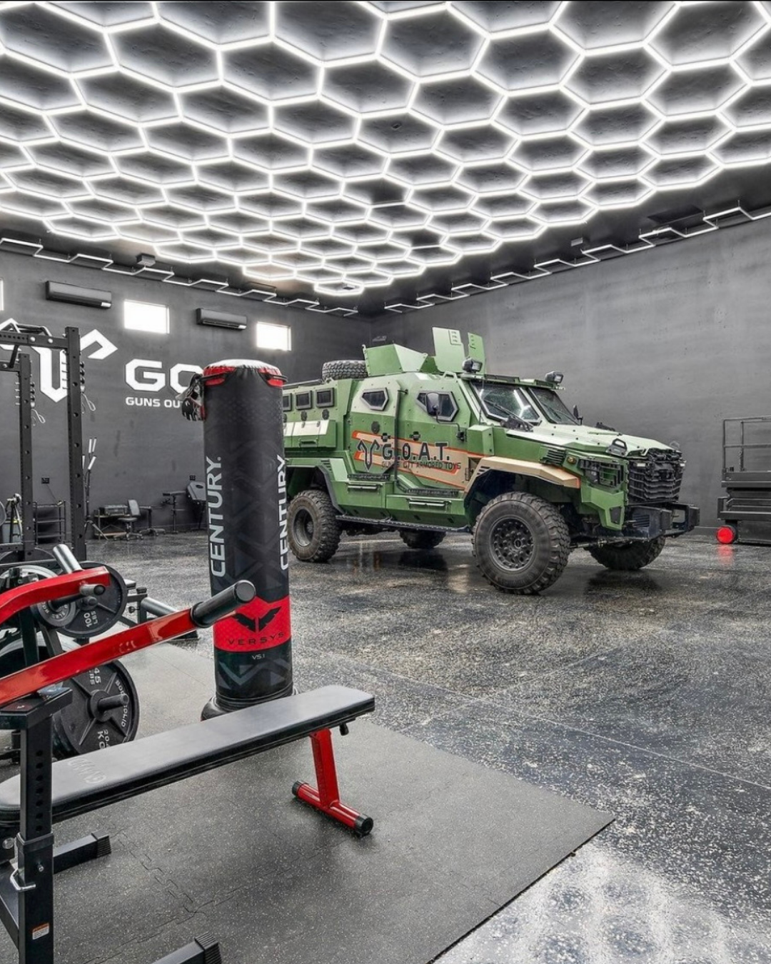 An industrial-style gym with a military armored vehicle parked inside. The gym has black walls with bright hexagonal ceiling lights, workout equipment including weights, a punching bag, and a bench.