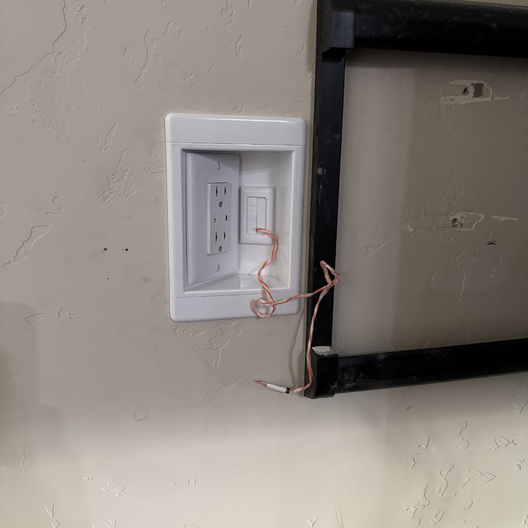 A wall outlet with two plugged-in electrical sockets, empty wire strands hanging, and a black metal frame nearby.