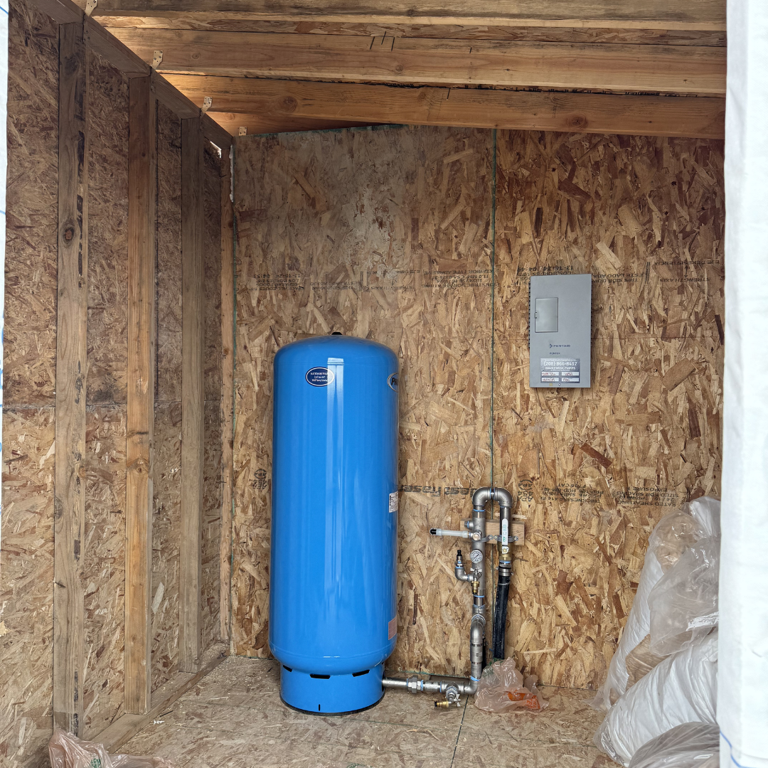 Inside a small unfinished room with wood walls and ceiling, featuring a large blue tank, plumbing pipes, and an electrical panel.